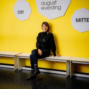 Prof. Dorte Eilers sitzend auf einer Bank im Akademietheater vor gelber Wand