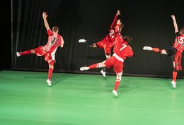Wir im Finale, vier Fußballspieler:innen sind von hinten zu sehen, alle mit dem linken Bein und Arm in der Luft, tanzend in der Formation