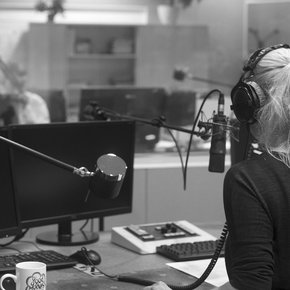 schwarz-weiß Bild blonde Frau sitzt mit Rücken zur Kamera in Radiostudio vor dem Mischpult