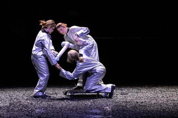 DARKNESS Produktionsfoto: Dunkler Hintergrund, drei Personen in silbernen Schutzanzügen angeleuchtet und in Aktion miteinander. Zwei stehend, eine Person knieend, dazwischen ein Roboterarm