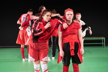 Wir im Finale, Fußballspieler auf dem Feld, eine der beiden Frauen im Vordergrund ist mit dem Arm auf ihre Nachbarin gestützt und hat ein Mikrophon in der Hand