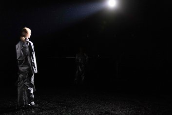 DARKNESS Produktionsfoto: Dunkle Bühne, ein Lichtspot gerichtet auf eine Person, die mit dem Rücken zum Betrachter steht, dahinter noch eine weitere Person kaum sichtbar