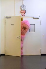QUERBALKEN: Person in rosa Flausch-Kostüm (Monsterchen) lugt aus Exit-Tür vor weißer Wand