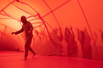 QUERBALKEN: Schemenhafte Gestalten außerhalb eines orangenen Tunnels aus dünnem Stoff, darin eine Person