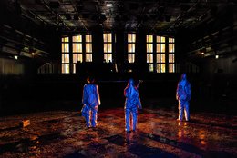 DARKNESS Produktionsfoto: Akademiestudiobühne, Fenster erleuchtet, auf der Bühne liegt brauner Staub, drei Personen stehen mit dem Rücken zum Betrachter, in blau ausgeleuchtet