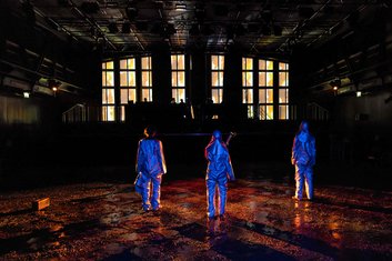 DARKNESS Produktionsfoto: Akademiestudiobühne, Fenster erleuchtet, auf der Bühne liegt brauner Staub, drei Personen stehen mit dem Rücken zum Betrachter, in blau ausgeleuchtet