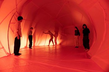 Querbalken: Fünf Personen stehen an den Wänden eines orangenen Tunnels aus dünnem Stoff