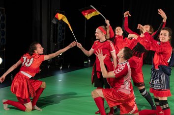 Wir im Finale, Fußballszene mit deutschen Fahnen, die Person links ist der Gruppe gegenüber und überreicht knieend eine Fahne