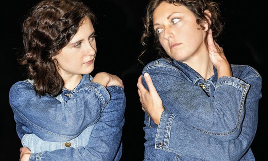 zwei Frauen in Jeansoveralls umarmen sich jeweils selbst und blicken sich dabei in die Augen