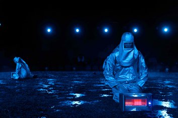 DARKNESS Produktionsfoto: Dunkle Szenerie, in blaues Licht getaucht, zwei Personen sitzen in silbernen Schutzanzügen auf der Bühne (wie Astronauten), ein rot leuchtendes technisches Gerät steht am Boden