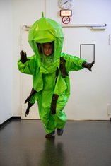 QUERBALKEN: eine Person in einem grünen Latex-Schutzanzug vor weißer Wand