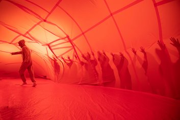 QUERBALKEN: Schemenhafte Gestalten in einem orangenen Tunnel aus dünnem Stoff