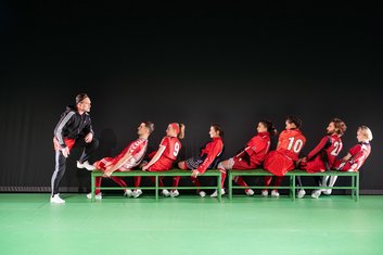 Wir im Finale, Fußballmannschaft sitzt auf Bank, Trainer links ihnen zugewandt, offenbar schreiend, die Mannschaft in roten Trikots lehnt sich von ihm weg