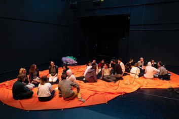 QUERBALKEN: 23 Schauspieler sitzen auf der schwarzen Bühne auf einem orangenen Tuch