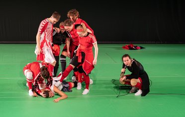 Wir im Finale, links eine Ansammlung von Fußballspielern im Kreis zusammen, rechts eine Dame in schwarzem Trikot mit Mikrophon