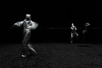 DARKNESS - Produktionsfoto: schwarzes Bild, schwarzer Boden, zwei Personen in silber gekleidet in Bewegung/im Tanz mit einem schwebenden Roboterarm